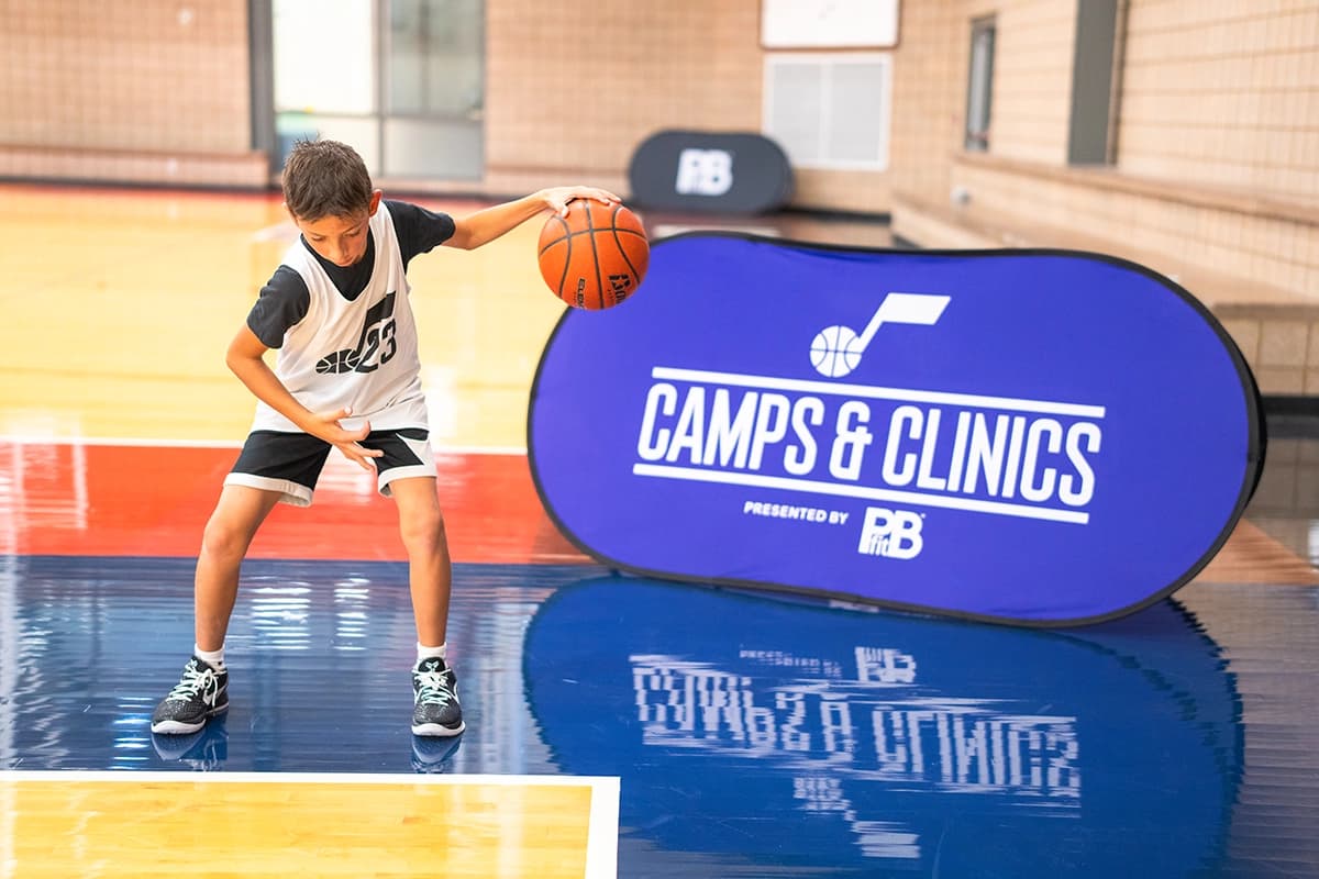 Young boy practicing basketball dribbling skills on an indoor court, wearing a jersey with the number 23. A blue banner in the background reads `Camps & Clinics presented by PBfit,` featuring a basketball and music note logo.