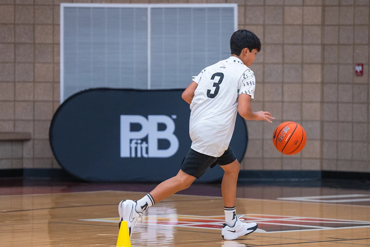 Young athlete practicing basketball dribbling drills on an indoor court, wearing a jersey with the number 3. A yellow cone is positioned on the floor, and a PBfit-branded banner is visible in the background.