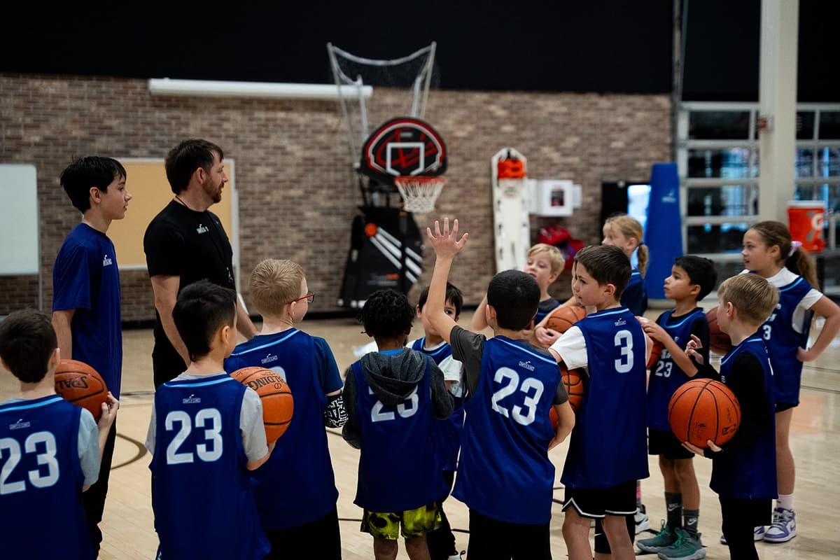 Group of young basketball players in blue jerseys gathered around a coach on an indoor court. One child raises their hand as others listen attentively, each holding a basketball. A basketball hoop and training equipment are visible in the background.